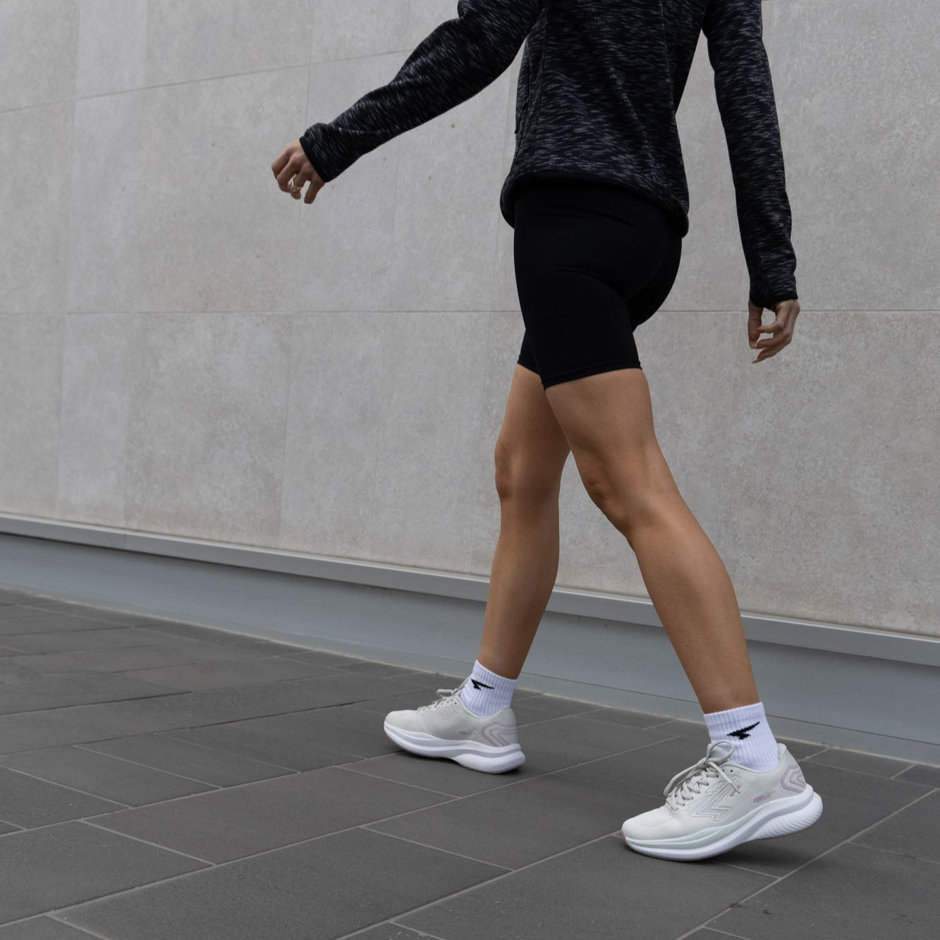 Person walking on a tiled pavement wearing black shorts, white sneakers, and dark long-sleeve top.