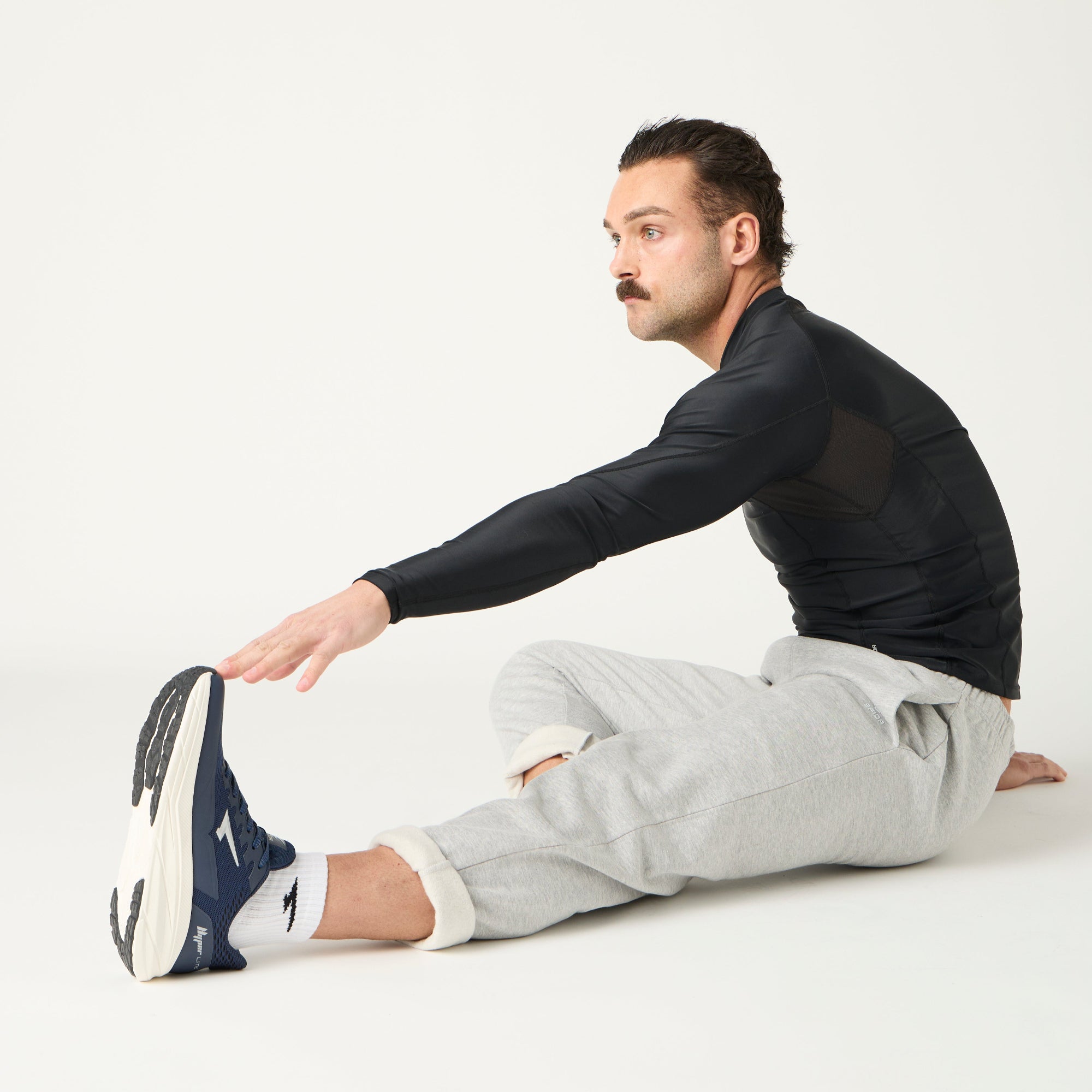 Man sitting on the floor wearing a black long-sleeve shirt, gray sweatpants, and navy sneakers on a white background