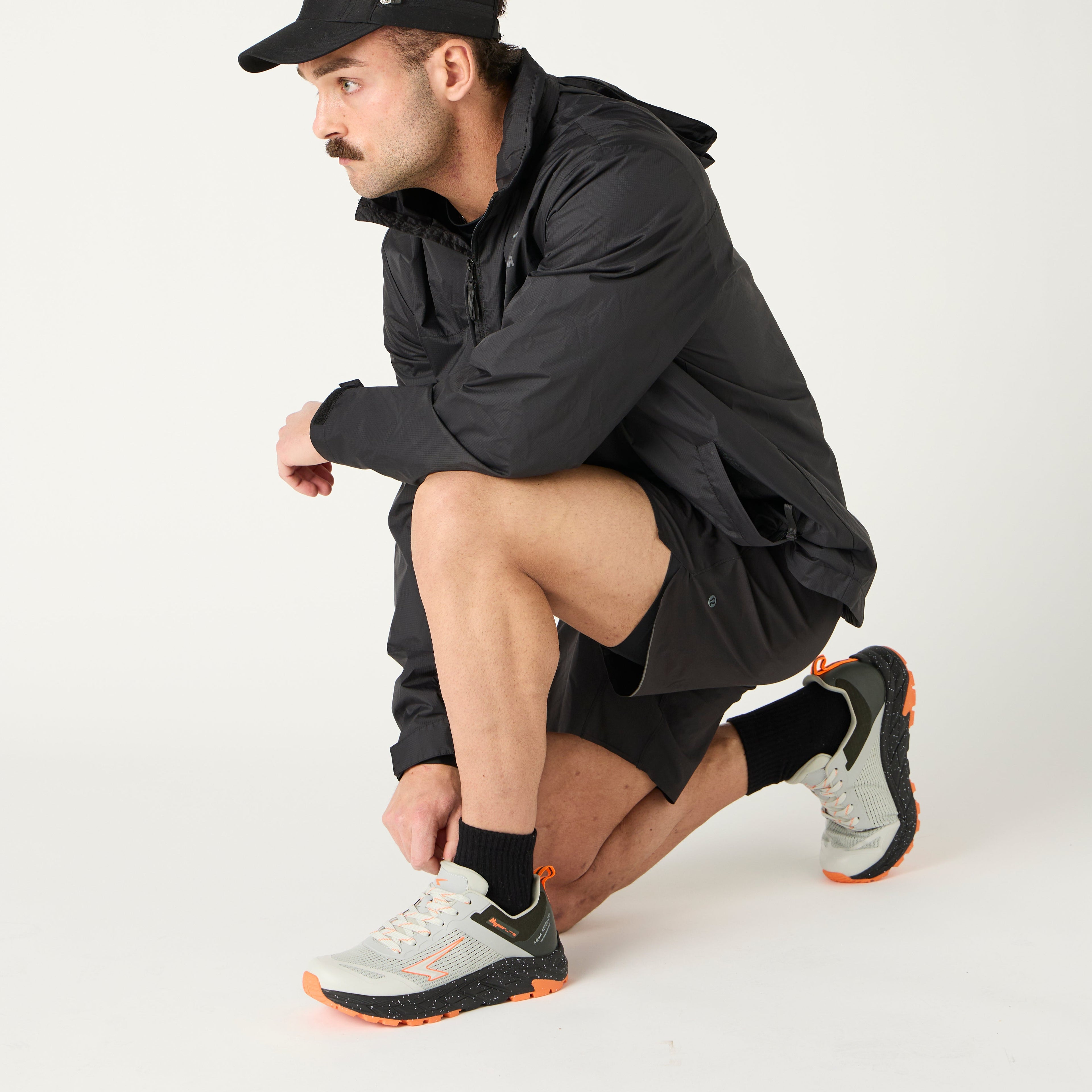 Man in black spray jacket with cap and trail shoes on a white background