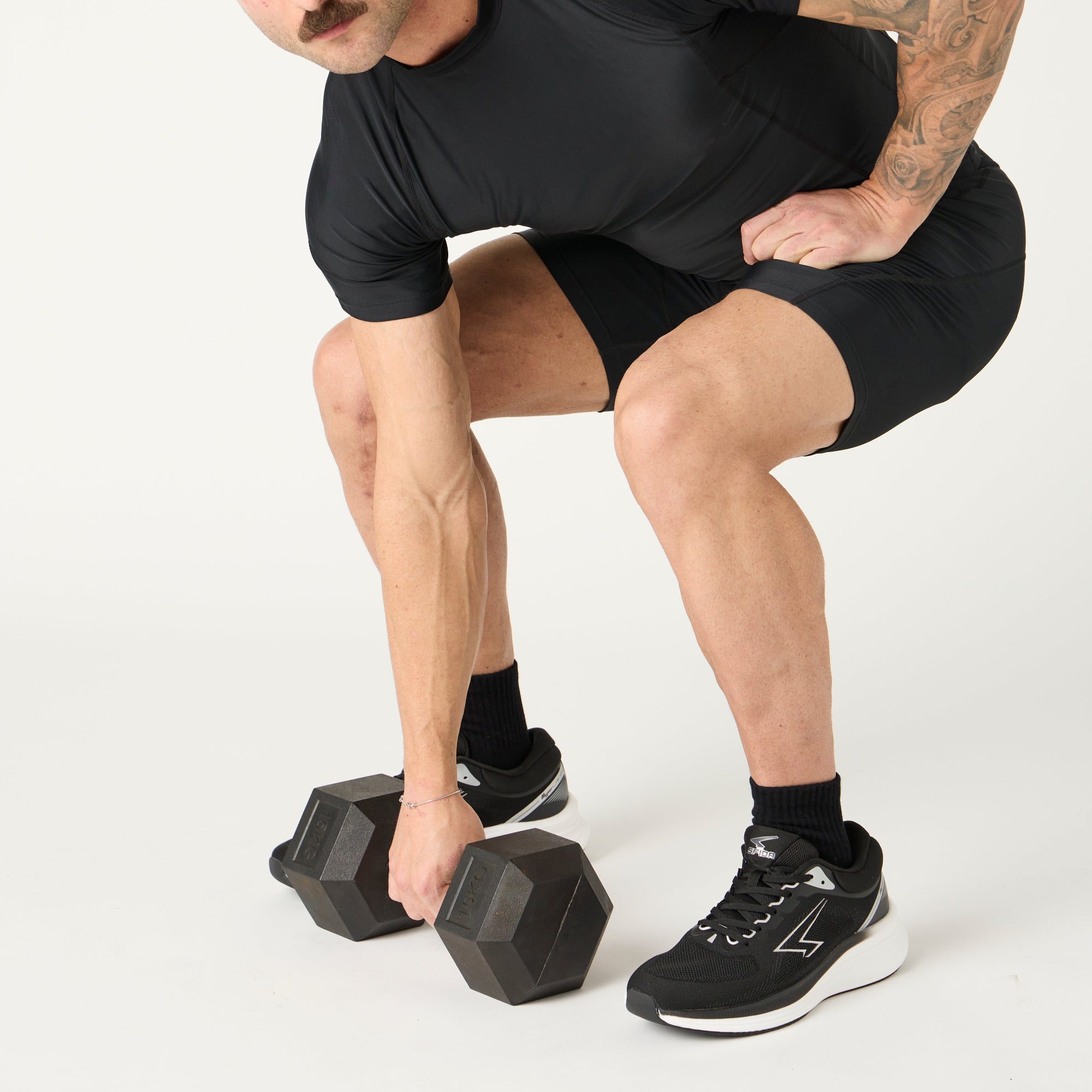 Man in black athletic compression wear and black runners performing a squat with dumbbells on a white background